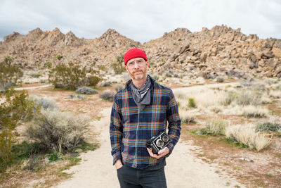 Man in beanie with beard and glasses by far off stone piles in desert