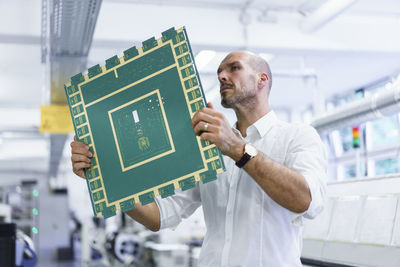 Confident mature male technician looking at large circuit board while examining in illuminated factory