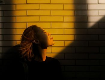 Side view of young woman sticking out tongue against wall during sunny day