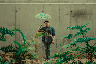 Portrait of man holding umbrella standing by wall amidst plants