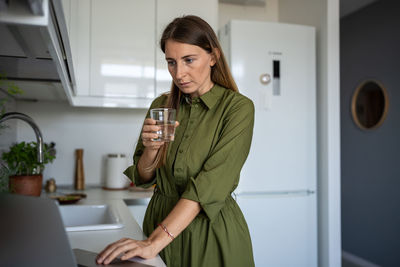 Focused woman standing on home kitchen with laptop, drinking water to calm down after working day.