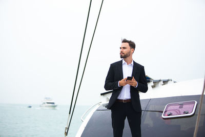 Businessman using phone while standing on boat in sea against clear sky