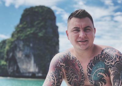 Portrait of shirtless young man with tattoo standing against sky at island