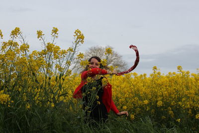 Portrait of cheerful young woman waving scarf while standing amidst yellow flowers blooming on field