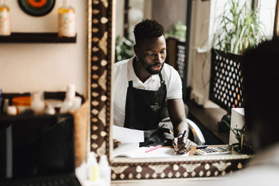 Reflection of male hairdresser calculating financial bill in barber shop