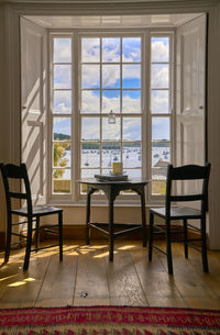 Empty chairs and a round table looking out through a large sunlit window to a view of boats at moor.