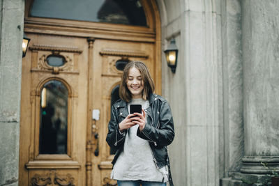 Full length of young woman using phone while standing on building