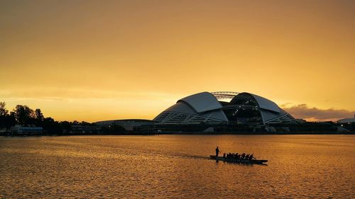 Silhouette boat sailing in river in front of singapore sports hub against orange sky during sunset