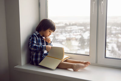 Boy child sits on the window of a house with a book in winter on a high floor
