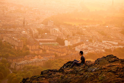 High angle view of man sitting on cliff against cityscape during sunset