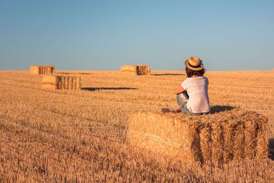 Rear view of woman sitting over hay bale on field against clear sky
