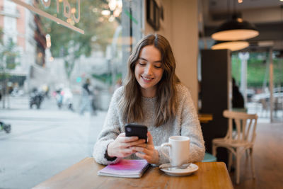 Content charming female sitting at table in cafe and browsing smartphone while smiling