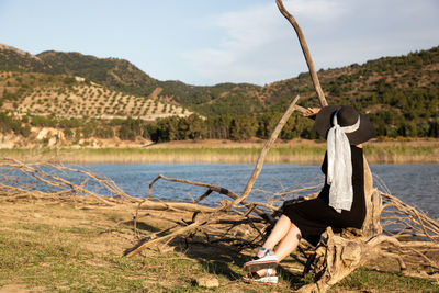 Woman sitting on fallen tree by lake against sky
