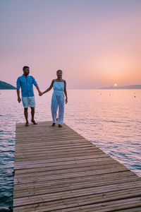 Rear view of men standing on sea against sky during sunset