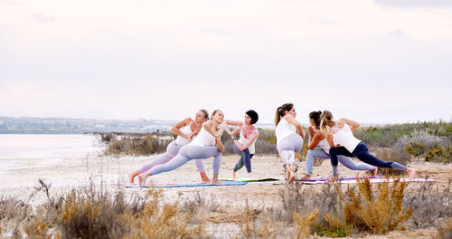 Female friends practicing yoga at beach against sky