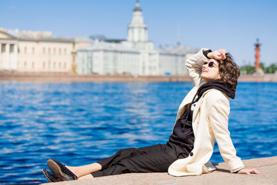 Girl is sitting on granite embankment. in white coat and sunglasses. city