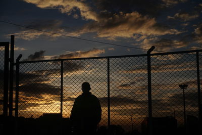 Silhouette man standing by fence against sky during sunset