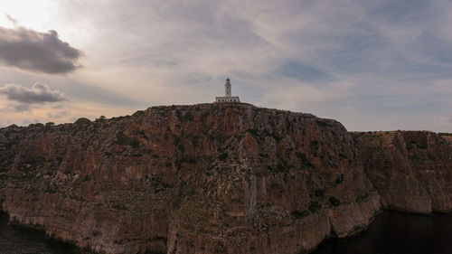 Low angle view of lighthouse on rocky mountain against cloudy sky