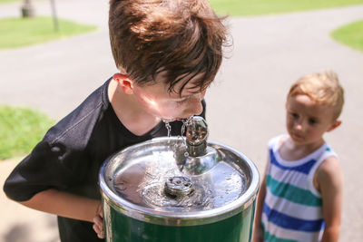 Cute boy looking at brother drinking water from container outdoors