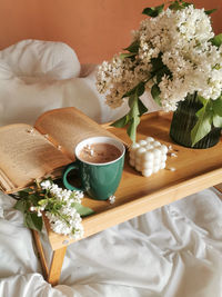 Folding wood table in bed with cup of coffe, open book, bubble cube candle and bunch of white lilac