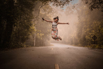 Full length of young woman with arms outstretched jumping on road amidst trees in forest