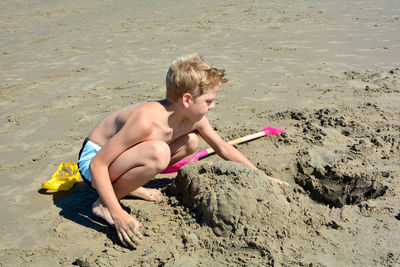 One blond boy with swimming trunks builds a sand castle in the sand on the beach