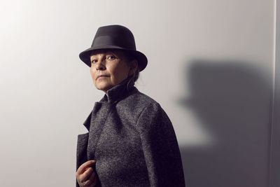 Fashionable older woman in black clothes and a hat stands against a white wall in the office