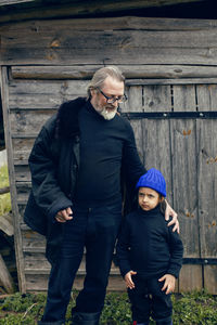 Grandson in a blue hat and boots with a grandfather in a sheepskin coat stand at a wooden house shed