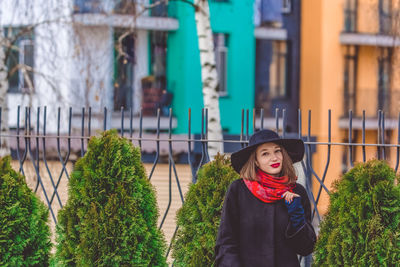 Portrait of woman wearing hat standing against fence in city