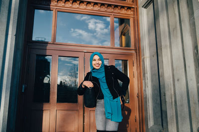 Low angle view of smiling young woman wearing hijab standing against door