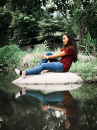 Side view of young woman sitting on rock in lake against trees