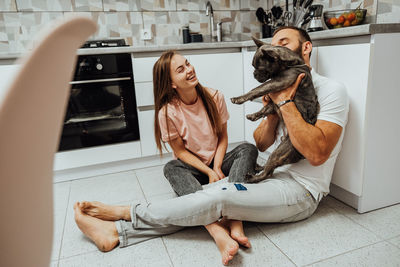 Cheerful woman and man having fun time at kitchen with their dog, french bulldog with family at home