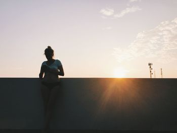 Woman using mobile phone while leaning on retaining wall against sky during sunset