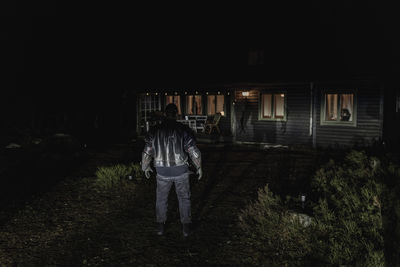 Rear view of demon standing in front of illuminated cabin at night