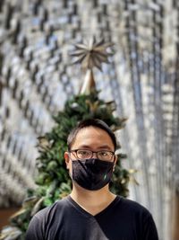 Portrait of young man wearing eyeglasses and face mask against christmas tree and skylight.