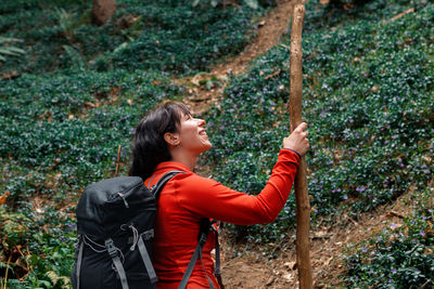 Side view of glad woman with backpack and stick admiring forest while travelling through coniferous woodland in weekend