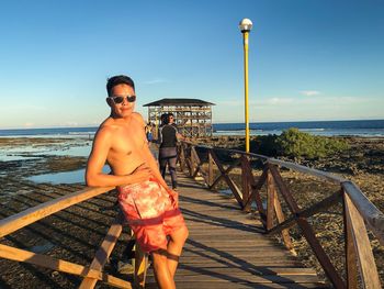 Portrait of shirtless man standing by railing against sea