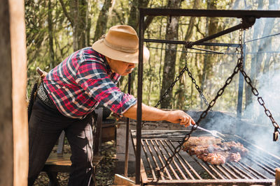 Side view of argentinian male in hat with fork adjusting fresh meat pieces on rack over flame in garden