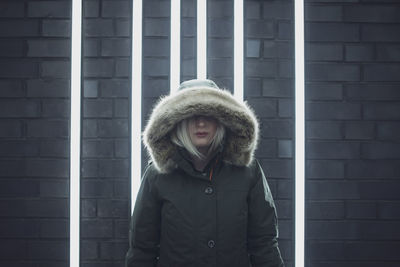 Young woman wearing fur hood while standing against illuminated brick wall