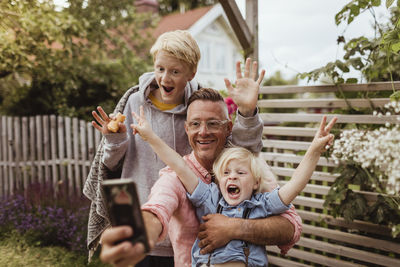 Cheerful father taking selfie with sons on smart phone in front yard at evening