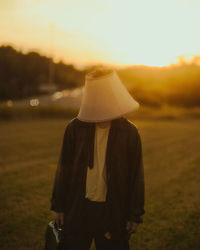 Front view of man wearing a lampshade on his head in front of a sunset.