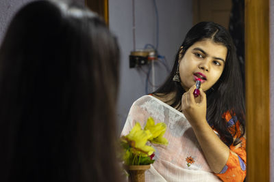 A beautiful indian woman in white saree applying lipstick in front of mirror with smiling face