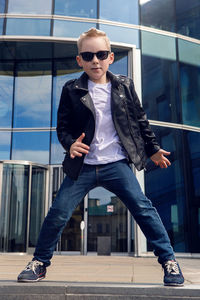 Baby boy in sunglasses and black leather jacket smiling on the background of the glass building