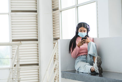 Young woman using mobile phone while sitting on window