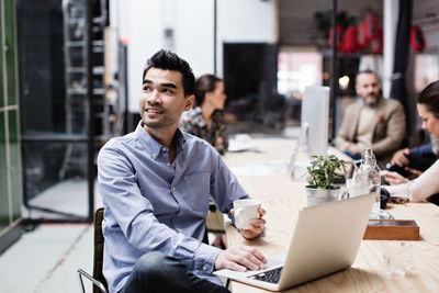 Smiling businessman having coffee and using laptop while sitting with colleagues at table