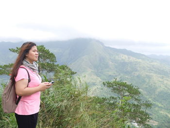 Side view of young woman with mobile phone standing on mountain against sky