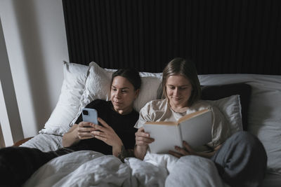 Female homosexual couple relaxing in bed, one woman using cell phone while another reading book