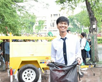 Portrait of smiling teenage boy holding garbage bag while standing by vehicle