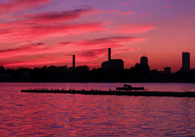 Silhouette factory by river against sky during sunset