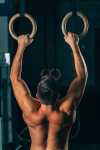 Rear view of shirtless male athlete exercising on gymnastics rings in gym